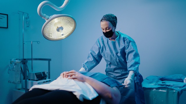 Dr. Beth Collins in sterile blue scrubs, adjusting surgical draping over a patient under an operating room light, demonstrating clinical excellence in plastic surgery.